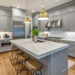Cocina gris a medida con isla central de mármol blanco, lámparas colgantes doradas y taburetes de madera