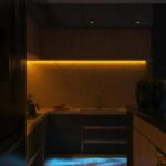 Cocina con iluminación LED en distintos circuitos: tira ambiental cálida bajo módulos altos y luces puntuales en techo